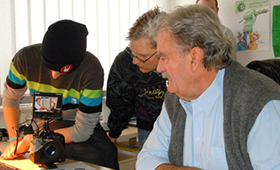 Jugendlicher hantiert an einem Tisch mit Kamera, Schauspieler Gerhard Olschewski blickt zu ihm hinber