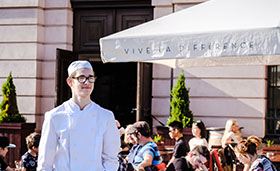 Ein junger Mann in der Kleidung eines Kochs steht im Auenbereich eines Restaurants. Bild: Gesellschaftsbilder.de | Andi Weiland