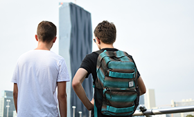 Zwei mnnliche Jugendliche mit Rucksack an einem Aussichtspunkt, im Hintergrund ein Hochhaus
