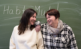 Zwei Frauen stehen vor einer Tafel und lächeln sich an, auf der Tafel steht "Habt Mut!" Zwei Frauen stehen vor einer Tafel und lächeln sich an, auf der Tafel steht "Habt Mut!"