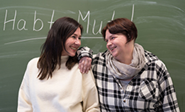Zwei Frauen stehen vor einer Tafel und l�cheln sich an, auf der Tafel steht "Habt Mut!"