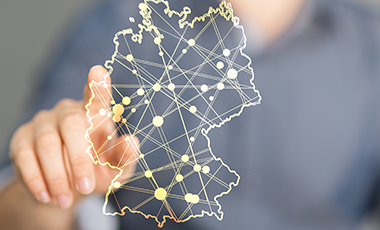 Im Vordergrund ist eine virtualisierte Deutschlandkarte zu sehen, im Hintergrund ein Mensch der mit dem Finger auf die Karte tippt. Im Vordergrund ist eine virtualisierte Deutschlandkarte zu sehen, im Hintergrund ein Mensch der mit dem Finger auf die Karte tippt.
