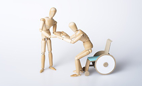 Symbolbild mit Maler-Gliederpuppen: Eine Figur hilft der anderen, aus dem Rollstuhl aufzustehen