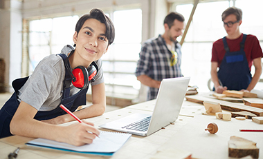 Weibliche Auszubildende steht an einem Tisch mit Laptop, im Hintergrund weitere männliche Auszubildende Weibliche Auszubildende steht an einem Tisch mit Laptop, im Hintergrund weitere männliche Auszubildende