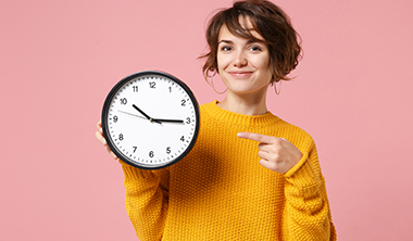 Junge Frau hält eine Wanduhr in der Hand und zeigt lächelnd darauf Junge Frau hält eine Wanduhr in der Hand und zeigt lächelnd darauf