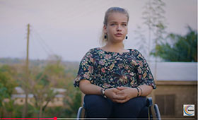 Im Vordergrund eine junge Frau im Rollstuhl. Im Hintergrund Bäume und niedrige Häuser. Im Vordergrund eine junge Frau im Rollstuhl. Im Hintergrund Bäume und niedrige Häuser.