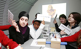 Im "Zeittunnel" des Projekts "komm auf Tour": Schülerinnen und Schüler sitzen an einem langen Tisch und halten Pappschilder mit "Vorher"- oder "Nachher"-Aufdruck hoch