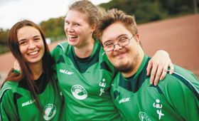 Drei lachende junge Menschen in  Trikots auf einem Fußballplatz