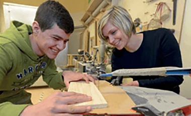 Auszubildender arbeitet an einem Holzbrett auf einer Werkbank, neben ihm steht eine Ausbilderin, beide lachen Auszubildender arbeitet an einem Holzbrett auf einer Werkbank, neben ihm steht eine Ausbilderin, beide lachen