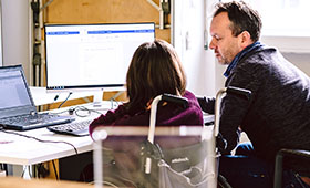 Eine Frau im Rollstuhl und ein Mann sitzen vor einem Tisch mit Laptop und Bildschirm. Bild: Andi Weiland | Gesellschaftsbilder.de