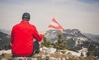 Mann sitzt auf einer Anh�he mit Blick auf ein Bergpanorama und h�lt eine �sterreichische Flagge in der Hand