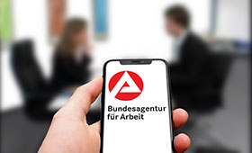 Smartphone, auf dem das BA-Logo zu sehen ist, im Hintergrund zwei Menschen im Gesprch