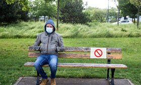 Junger Mann mit Maske sitzt auf einer Parkbank, auf der auf einer Seite ein "Verboten"-Schild angebracht ist.