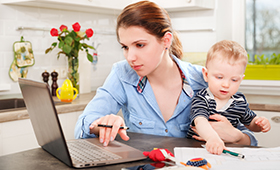 Junge Mutter vor Laptop mit Kleinkind auf dem Arm