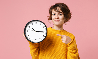 Junge Frau hält eine Wanduhr in der Hand und zeigt lächelnd darauf Junge Frau hält eine Wanduhr in der Hand und zeigt lächelnd darauf