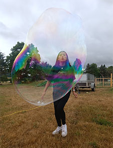 Eine junge Frau steht auf einer Wiese und macht eine riesige Seifenblase. Bild: ProLand