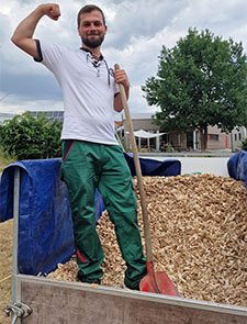 Ein junger Mann steht auf einem Wagen mit Mulch. In einer Hand hlt er eine Schaufel. Den anderen Arm hebt er, um die Muskeln zu zeigen.