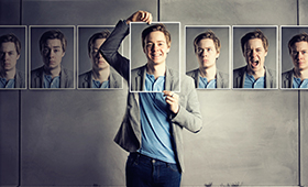 Junger Mann hält ein Foto seiner selbst vor sich hin, links und rechts an den Wänden sind auf gleicher Höhe weitere Fotos von ihm mit unterschiedlichen Gesichtsausdrücken