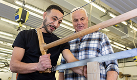 Auszubildender sgt einen Holzblock, neben ihm steht ein Ausbilder