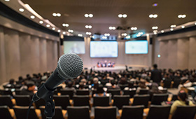 Blick aus der letzten Publikumsreihe auf das Podium eines Tagungsraums, im Vordergrund ein Mikrofon