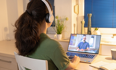 Rückansicht einer Frau mit Kopfhörern, am Schreibtisch vorm Laptop sitzend, auf dem Bildschirm ein Mann ebenfalls an einem Tisch Rückansicht einer Frau mit Kopfhörern, am Schreibtisch vorm Laptop sitzend, auf dem Bildschirm ein Mann ebenfalls an einem Tisch