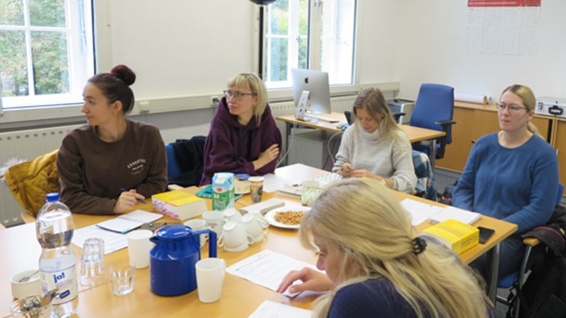 Eine Gruppe von Frauen sitzt um einen Tisch, auf dem Seminarunterlagen liegen. Alle schauen auf eine Leinwand. Bild: TH Kln