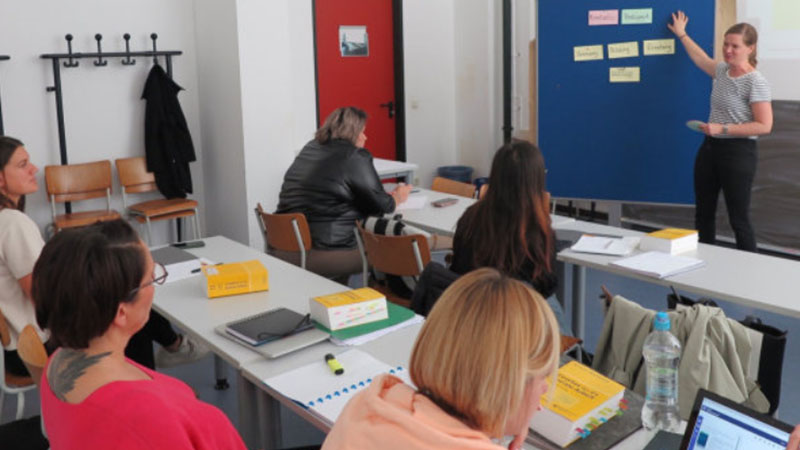Eine Gruppe in einem Seminarraum. Die Dozentin zeigt etwas an einer Pinnwand. Bild: TH Kln