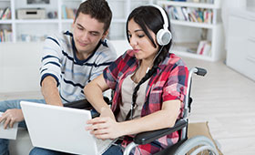 Junge Frau im Rollstuhl und neben ihr sitzender junger Mann schauen gemeinsam auf den Bildschirm eines Laptops