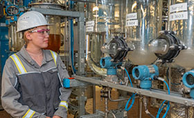 Junge Auszubildende mit Schutzhelm vor einer Industrieanlage mit Leitungen und Rohren