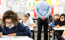 Schlerinnen schreiben einen Test. Ein Lehrer, mit dem Rcken zur Kamera, beaufsichtigt sie. Bild: rawpixel.com | Adobe Stock