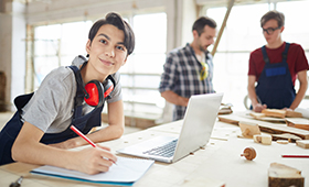 Weibliche Auszubildende steht an einem Tisch mit Laptop, im Hintergrund weitere mnnliche Auszubildende