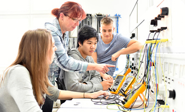 Eine kleine Gruppe von Jugendlichen vor einem Pult mit elektronischen Anlagen, stehend daneben eine Ausbilderin Eine kleine Gruppe von Jugendlichen vor einem Pult mit elektronischen Anlagen, stehend daneben eine Ausbilderin