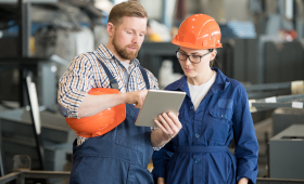 Ein Mann hlt ein Tablet in der Hand und erklrt einer Auszubildenden etwas. Die Auszubildende trgt einen orangenen Schutzhelm.