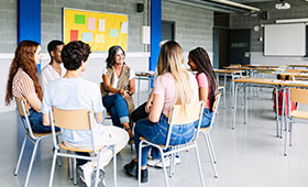 Junge Menschen sitzen mit ihrer Lehrerin in einem Stuhlkreis in einem Klassenzimmer. Bild: Xavier Lorenzo | Adobe Stock
