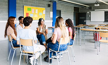 Junge Menschen sitzen mit ihrer Lehrerin in einem Stuhlkreis in einem Klassenzimmer.