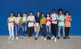10 Schlerinnen und Schler verschiedener ethnischer Herkunft stehen mit Schulunterlagen in der Hand nebeneinander vor einer blauen Wand.