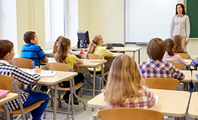Jungen und Mdchen in einer Schulklasse, im Hintergrund steht die Lehrerin an der Schultafel