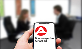 Smartphone, auf dem das Logo der Bundesagentur fr Arbeit zu sehen ist, im Hintergrund zwei Menschen im Gesprch