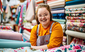 Lachende junge Frau mit Down-Syndrom inmitten von bunten Stoffballen in einem Stoffgeschft