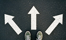 Ein paar Fe in Sneakers vor drei Pfeilen auf dunklem Asphalt, die in verschiedene Richtungen weisen