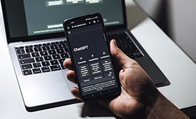Eine Hand h�lt ein Smartphone, auf dem Chat GPT aufgerufen ist, dahinter ein Laptop