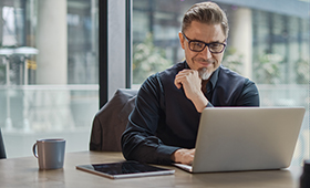 Junger Mann sitzt am Schreibtisch und blickt l�chelnd auf ein Laptop