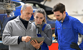 Ausbilder erl�utert Auszubildender und Auszubildendem etwas mit dem Laptop in der Hand, im Hintergrund ein Auto auf einer Hebeb�hne