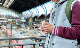 Jugendlicher mit Handy in der Hand, im Hintergrund die Bahnsteige eines gro�en Bahnhofs