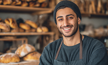 Person mit Migrationshintergrund tr�gt eine B�ckersch�rze und steht vor einem Regal Br�tchen.