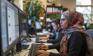 Eine junge Frau mit Kopftuch sitzt vor einem PC. Im Hintergrund sind weitere Personen zu erkennen, die am PC arbeiten.