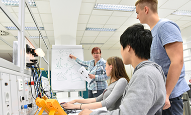 Drei Jugendliche lernen an elektronischen Geräten in einem Industriebetrieb. Eine Lehrerin steht an einem Whiteboard und erklärt etwas. Drei Jugendliche lernen an elektronischen Geräten in einem Industriebetrieb. Eine Lehrerin steht an einem Whiteboard und erklärt etwas.