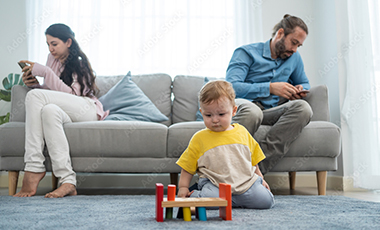 Mutter und Vater sitzen auf einem Sofa und blicken auf ihre Smartphones, vor ihnen auf dem Teppich spielt ihr kleiner Junge mit Spielzeug Mutter und Vater sitzen auf einem Sofa und blicken auf ihre Smartphones, vor ihnen auf dem Teppich spielt ihr kleiner Junge mit Spielzeug