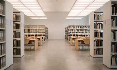 Langer Gang in einer modernen Bibliothek, hell beleuchtet. Weiße Regale rechts und links sowie Arbeitsplätze im Hintergrund. Langer Gang in einer modernen Bibliothek, hell beleuchtet. Weiße Regale rechts und links sowie Arbeitsplätze im Hintergrund.